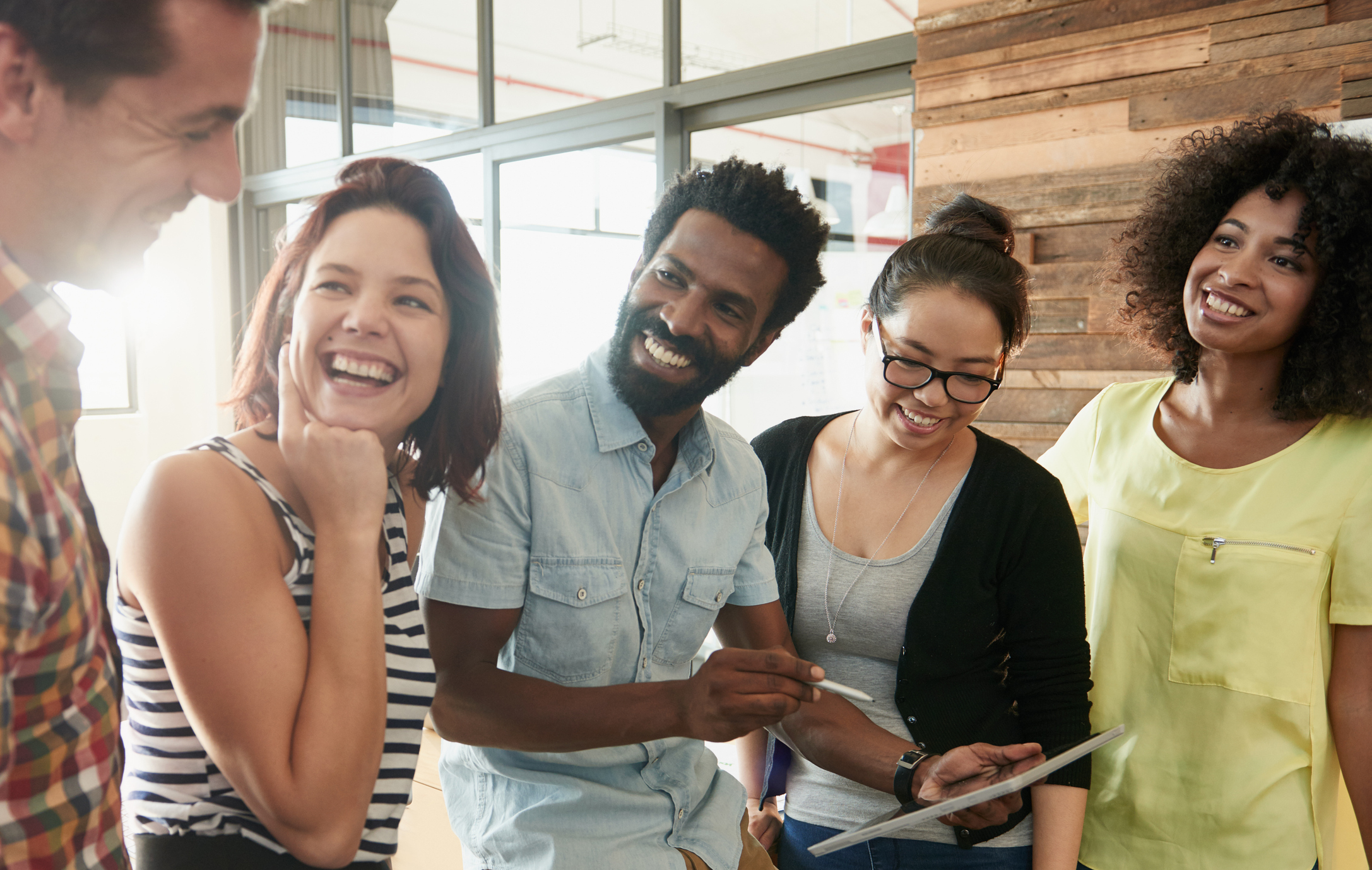 A photo of a diverse group of men and women looking together at a tablet and smiling in amusement